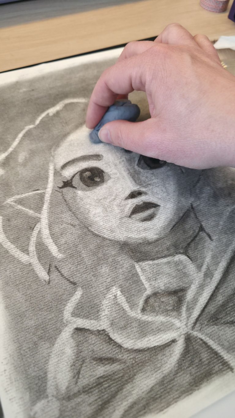 Gros plan d'une main d'artiste utilisant une gomme mie de pain sur le front d'un portrait de jeune fille (Zelda) dessinée au fusain.