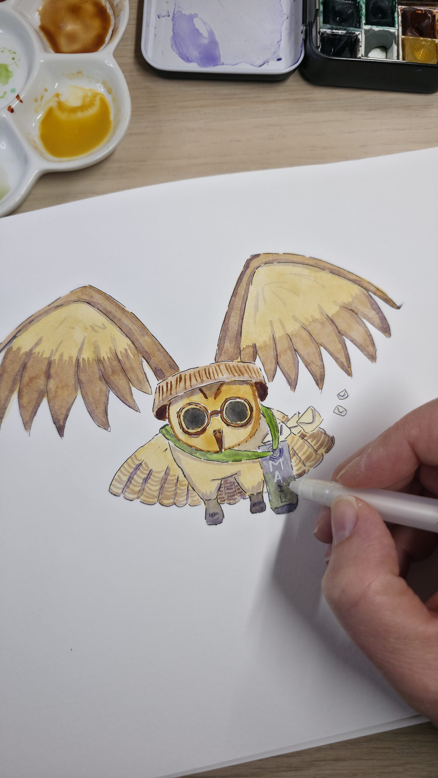 Photographie d'une main d'artiste utilisant un stylo blanc pour ajouter des détails sur une illustration à l'aquarelle représentant un petit hibou facteur avec des lunettes et une sacoche de courrier.