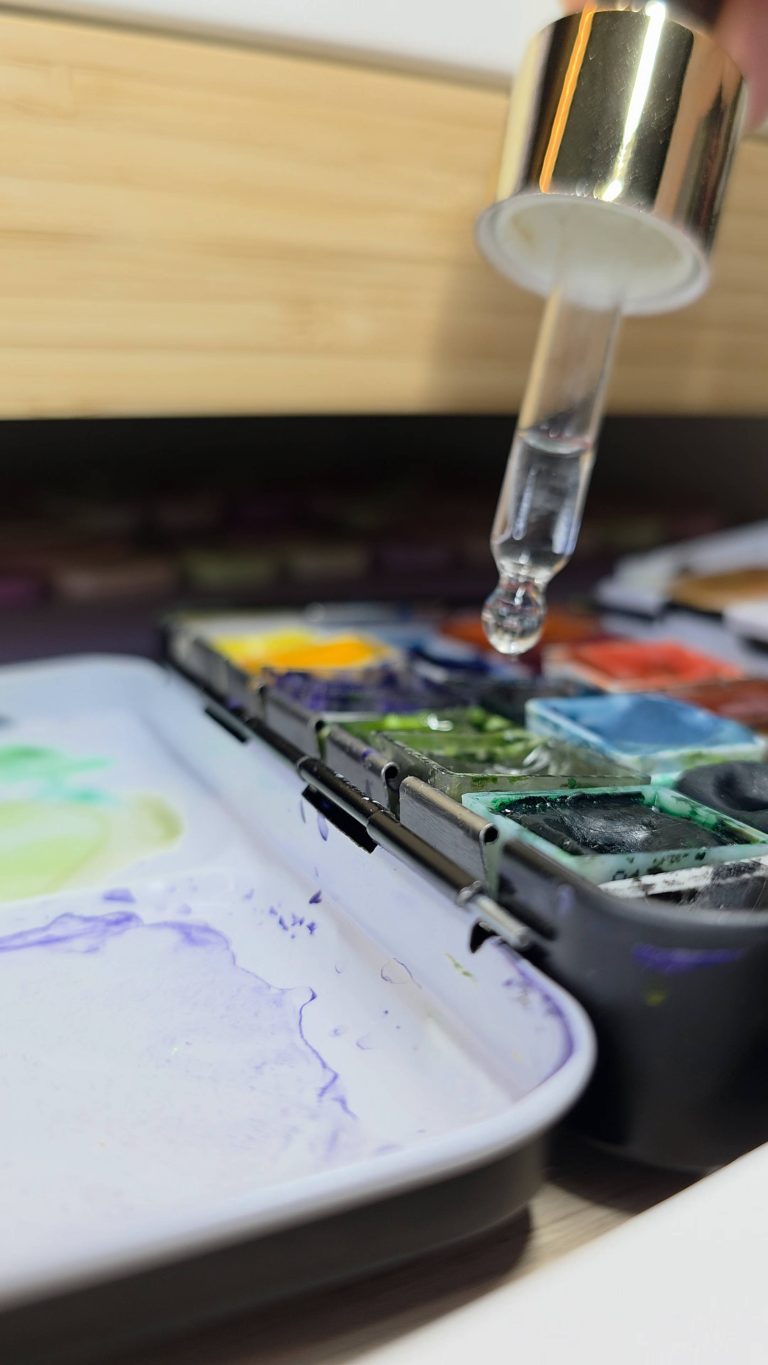Photographie en gros plan d'une pipette en verre déposant une goutte d'eau dans un godet de peinture sur une palette d'aquarelle.